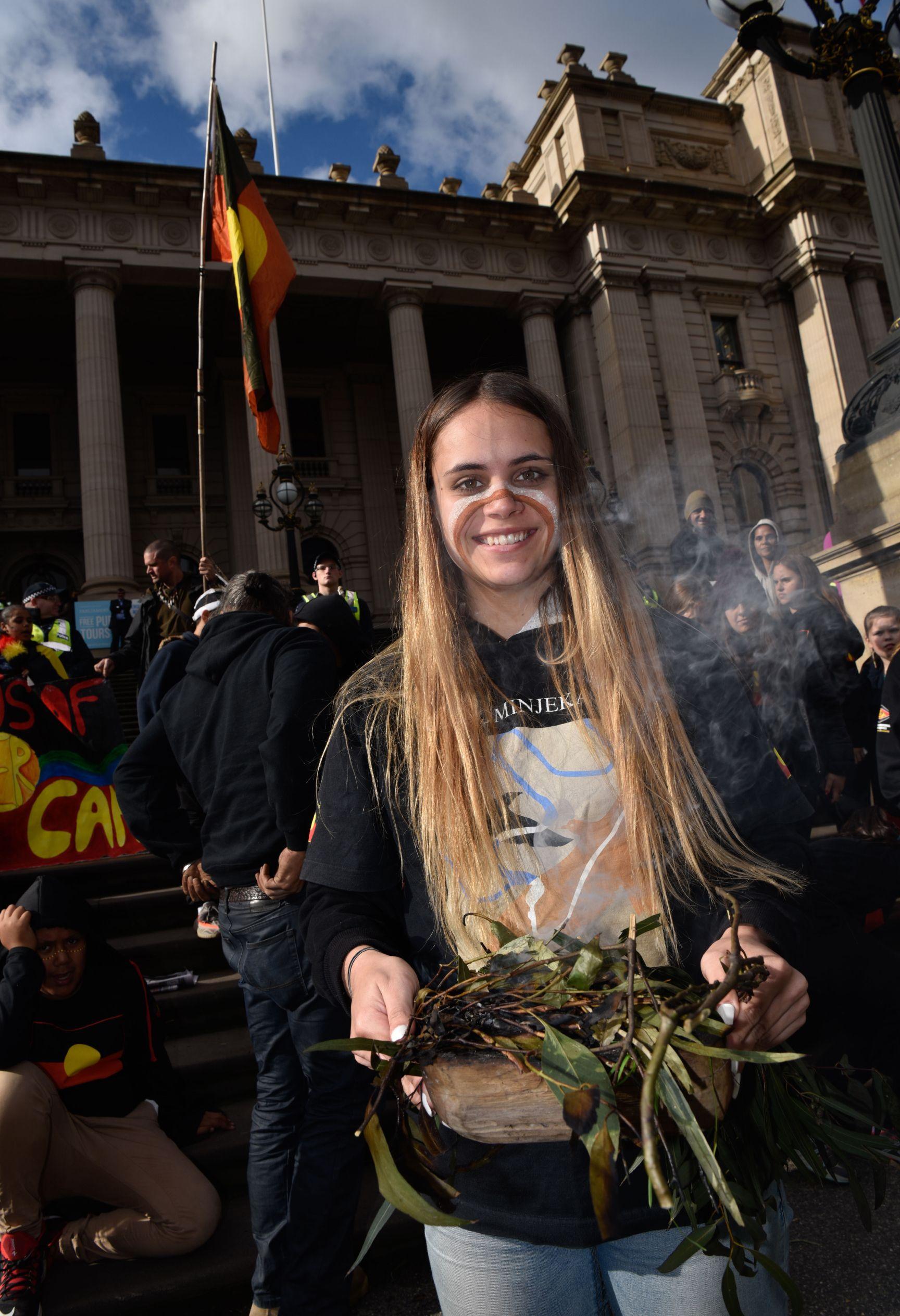 A young indigenous woman wearing traditional face paint and a shirt that reads "Wominjeka" stands in front of a crowd of people outside Melbourne's Parliament House. She is holding materials for a smoking ceremony.