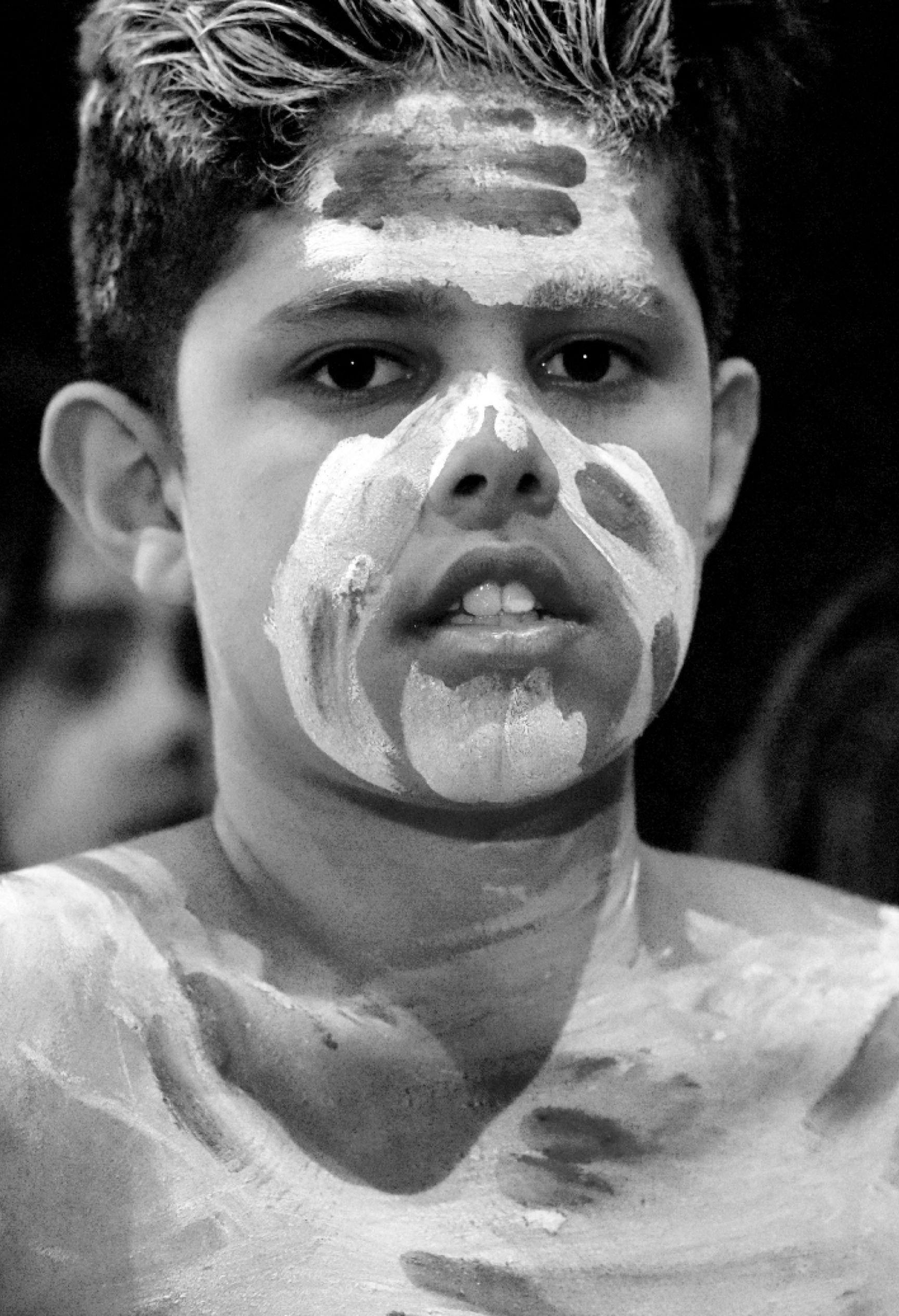 A black and white photo of a young indigenous boy wearing traditional face and body paint.